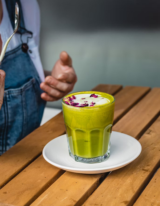 A close-up, slightly high-angle shot of a vibrant green matcha latte in a clear, fluted glass, topped with a few dried rose petals. The glass sits on a white saucer atop a wooden slatted table, with a person wearing denim overalls partially visible in the background, making this a relaxing cafe moment.