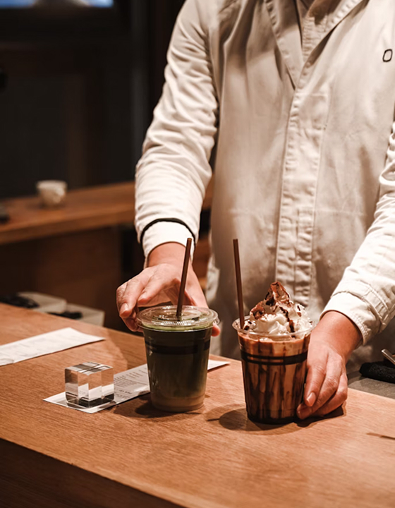 A close-up shot of a person in a light-colored apron carefully placing two drinks on a wooden bar counter. One is a vibrant iced matcha beverage and the other is a dark iced coffee, capturing a quiet and relaxing cafe ritual.