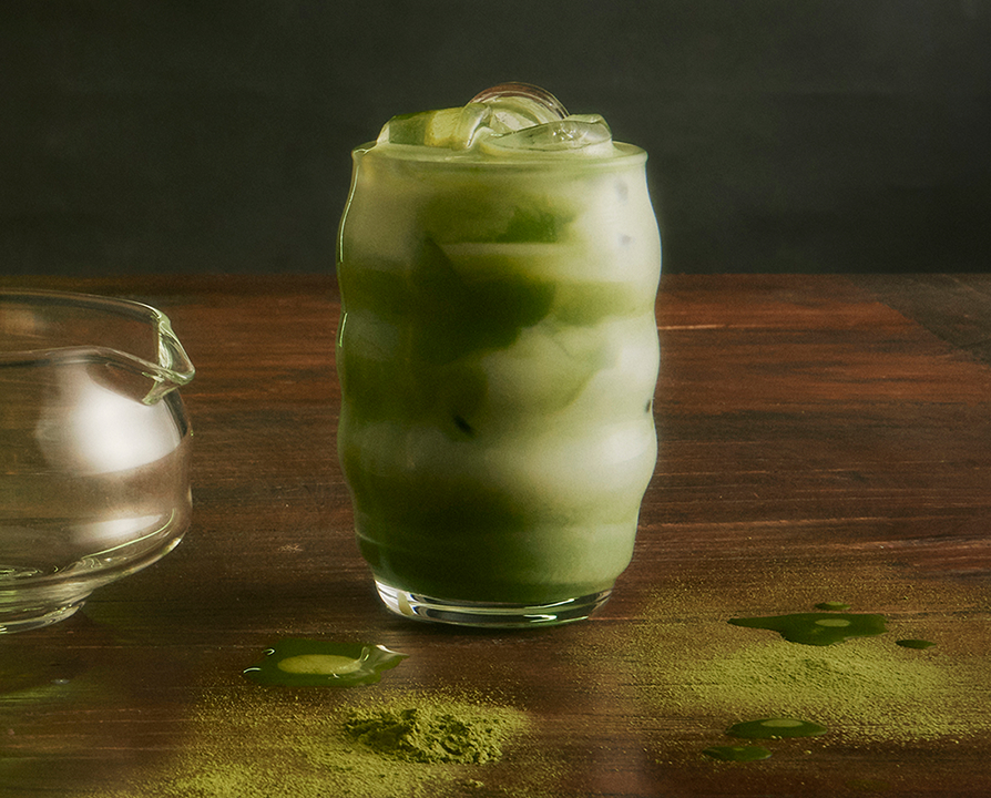 Glass of iced matcha latte with swirls of green tea and milk on a wooden surface with matcha powder and drops scattered around.