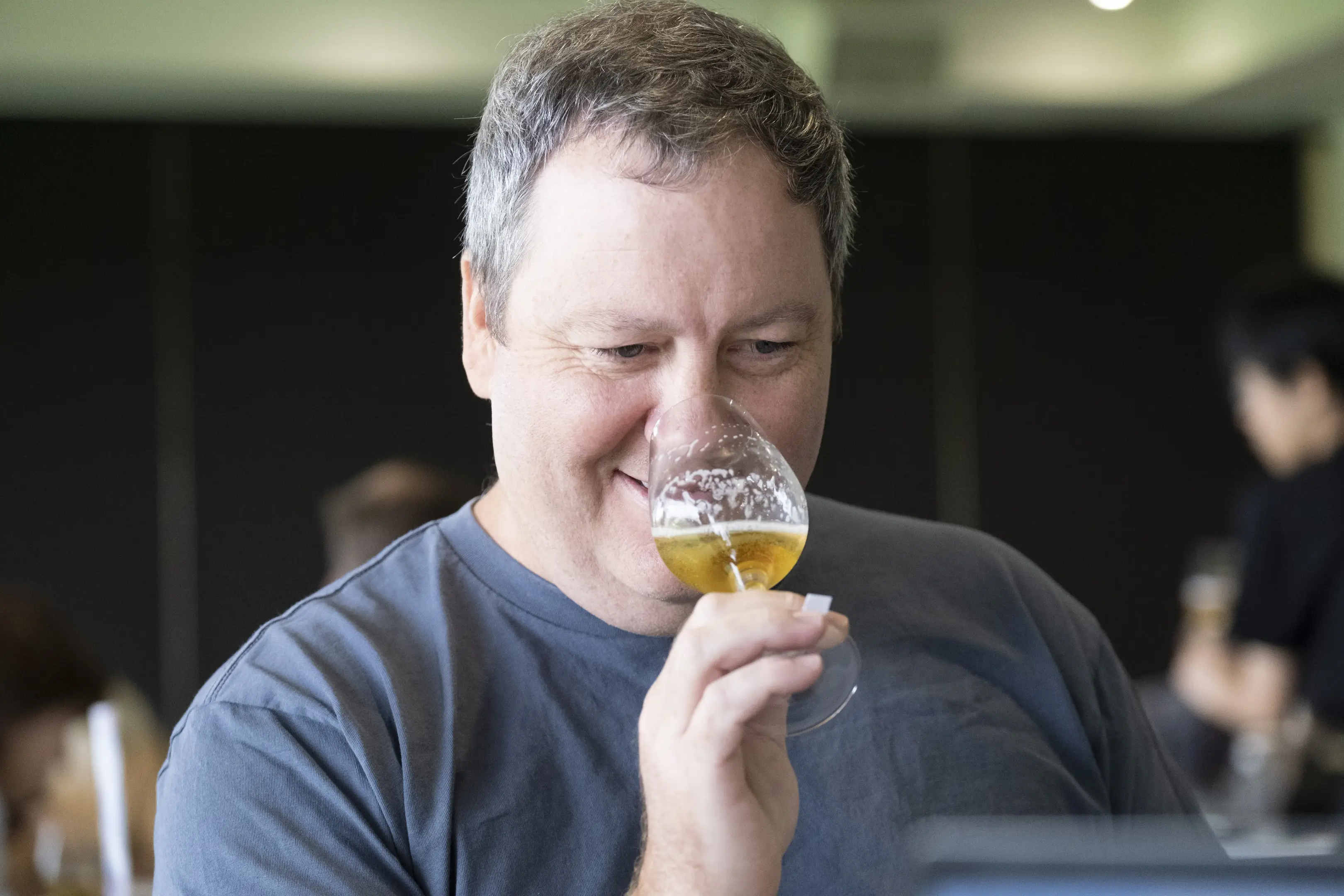A man with gray hair and a gray shirt is smiling while sniffing a glass of beer in an indoor setting.