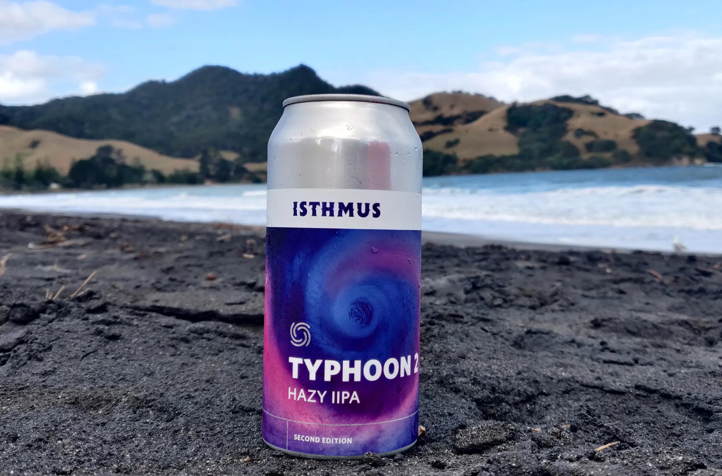 A can of Typhoon Hazy IPA beer stands on dark sand with the ocean and hilly landscape in the background under a partly cloudy sky.