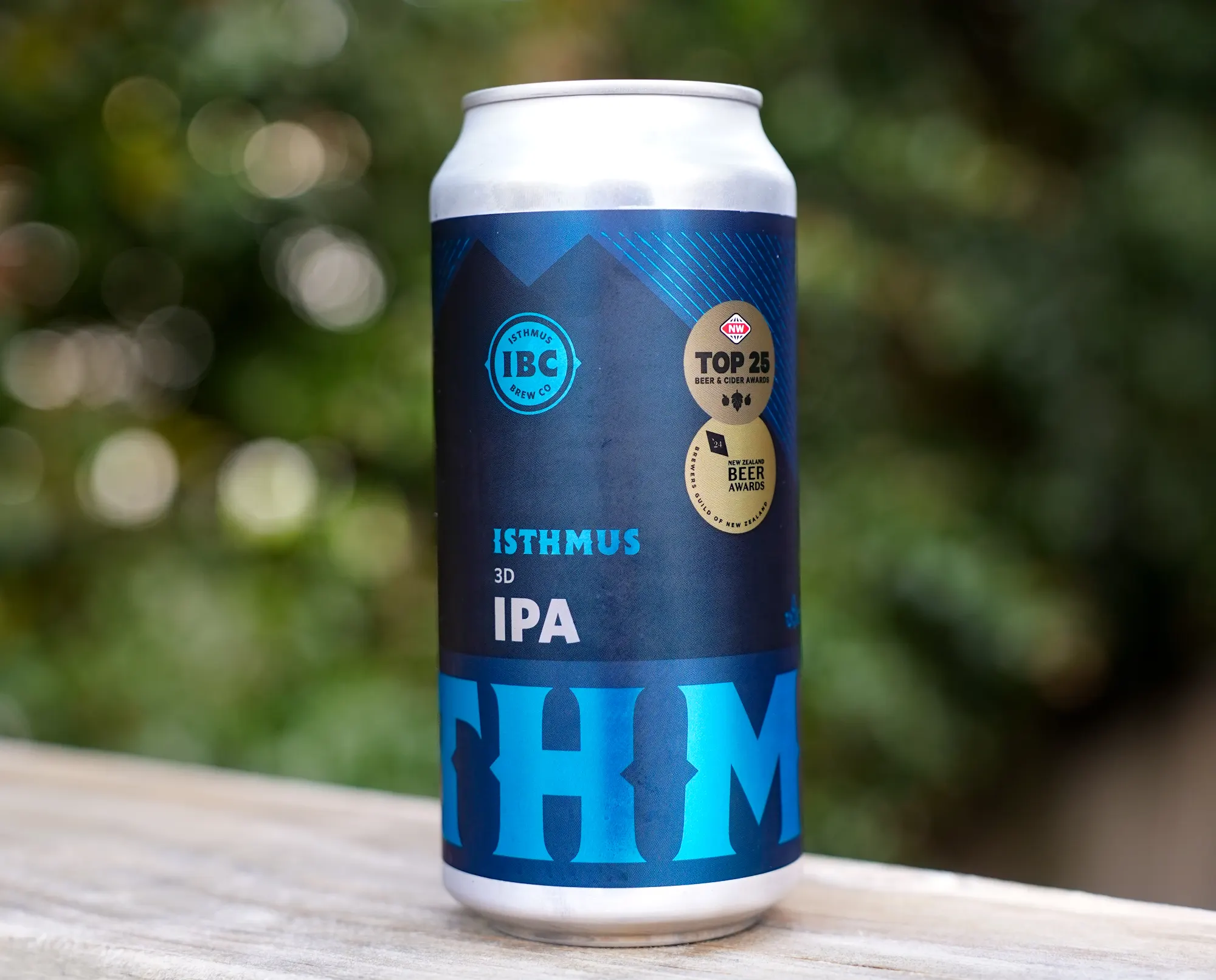 A can of Isthmus IPA beer sits on a wooden post in front of grassy sand dunes, with a cloudy sky and the ocean visible in the background.