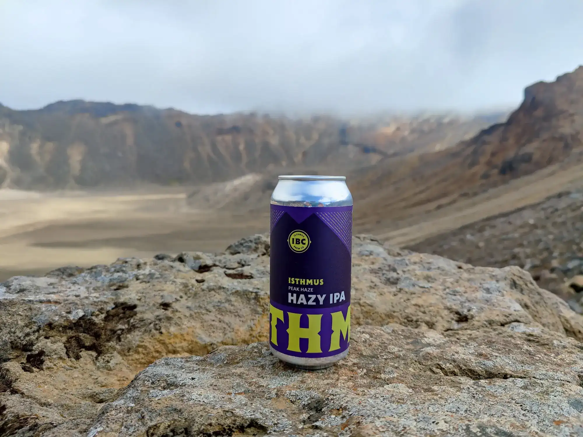 A can of hazy IPA beer sits on a rocky surface, with a misty, mountainous landscape and cloudy sky in the background.