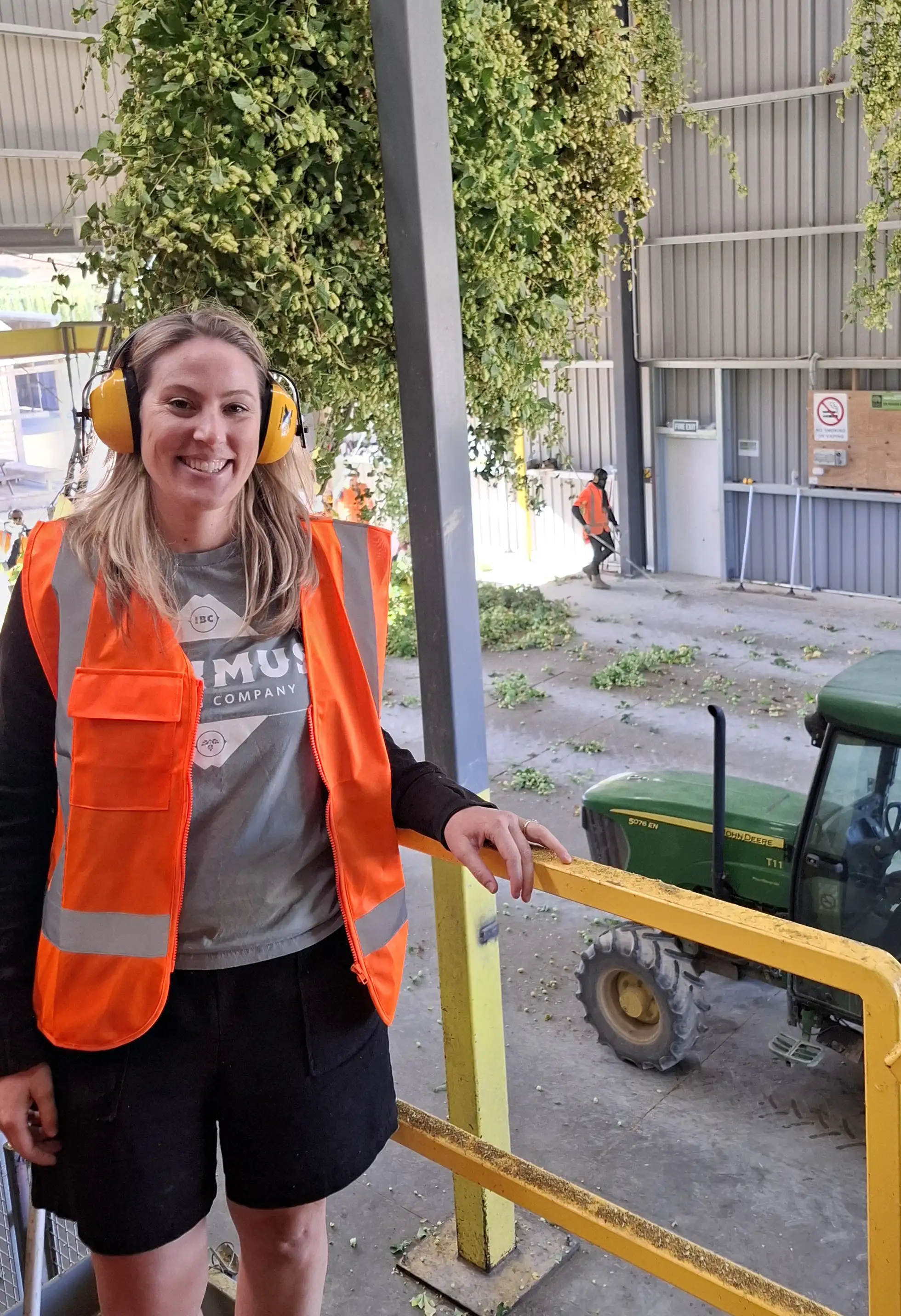 A person wearing an orange safety vest, ear protection, and black shorts stands next to a yellow railing inside an industrial facility, with a green tractor and hanging plants visible in the background.