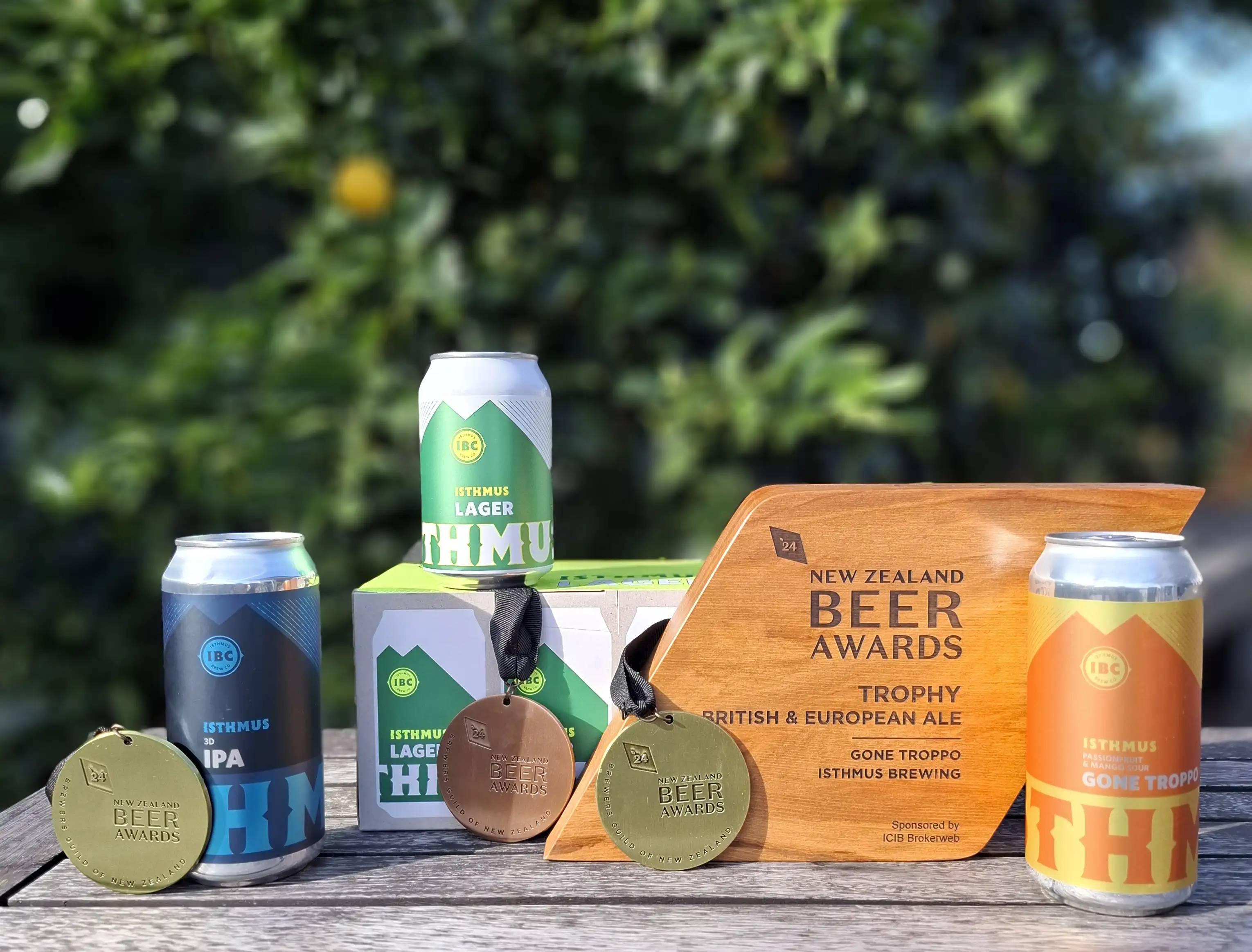Three cans of Isthmus Brewing Co. beer, two medals, and a NZ Beer Awards trophy are displayed on a wooden surface outdoors, with greenery blurred in the background.