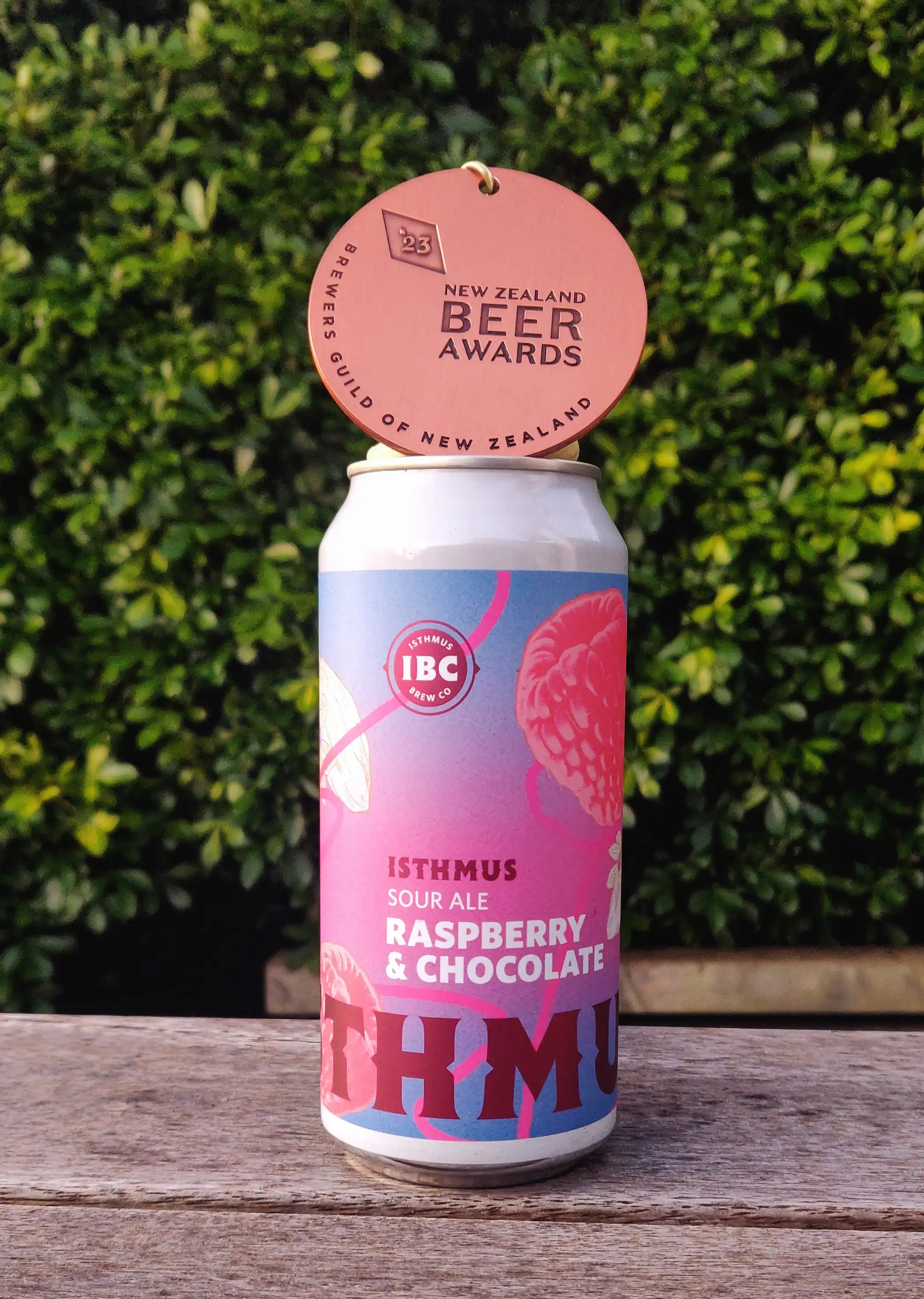 A can of beer sits on a wooden surface outdoors, with greenery in the background and a round award on top