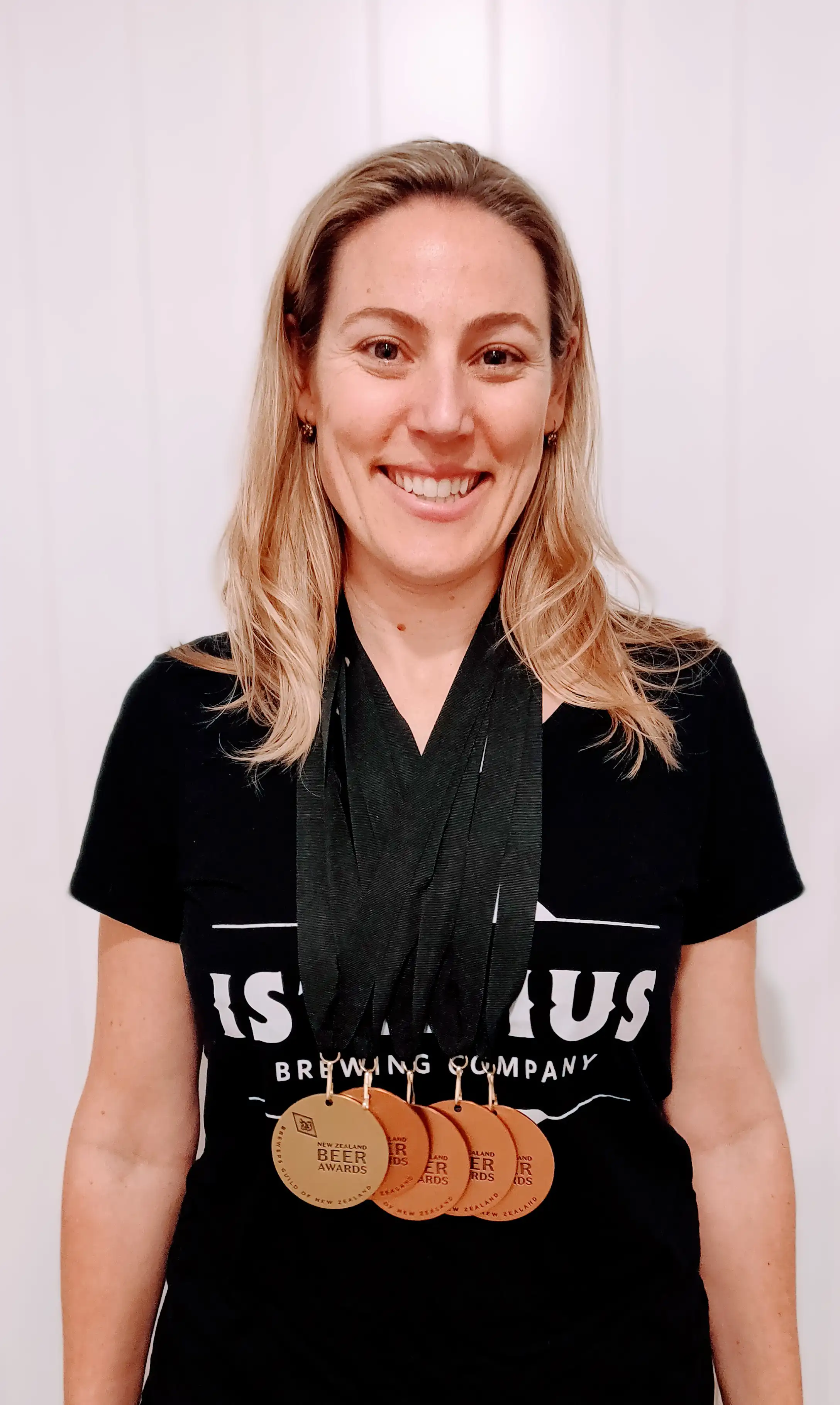 A woman with blonde hair, wearing a black t-shirt, smiles at the camera while displaying several medals around her neck.