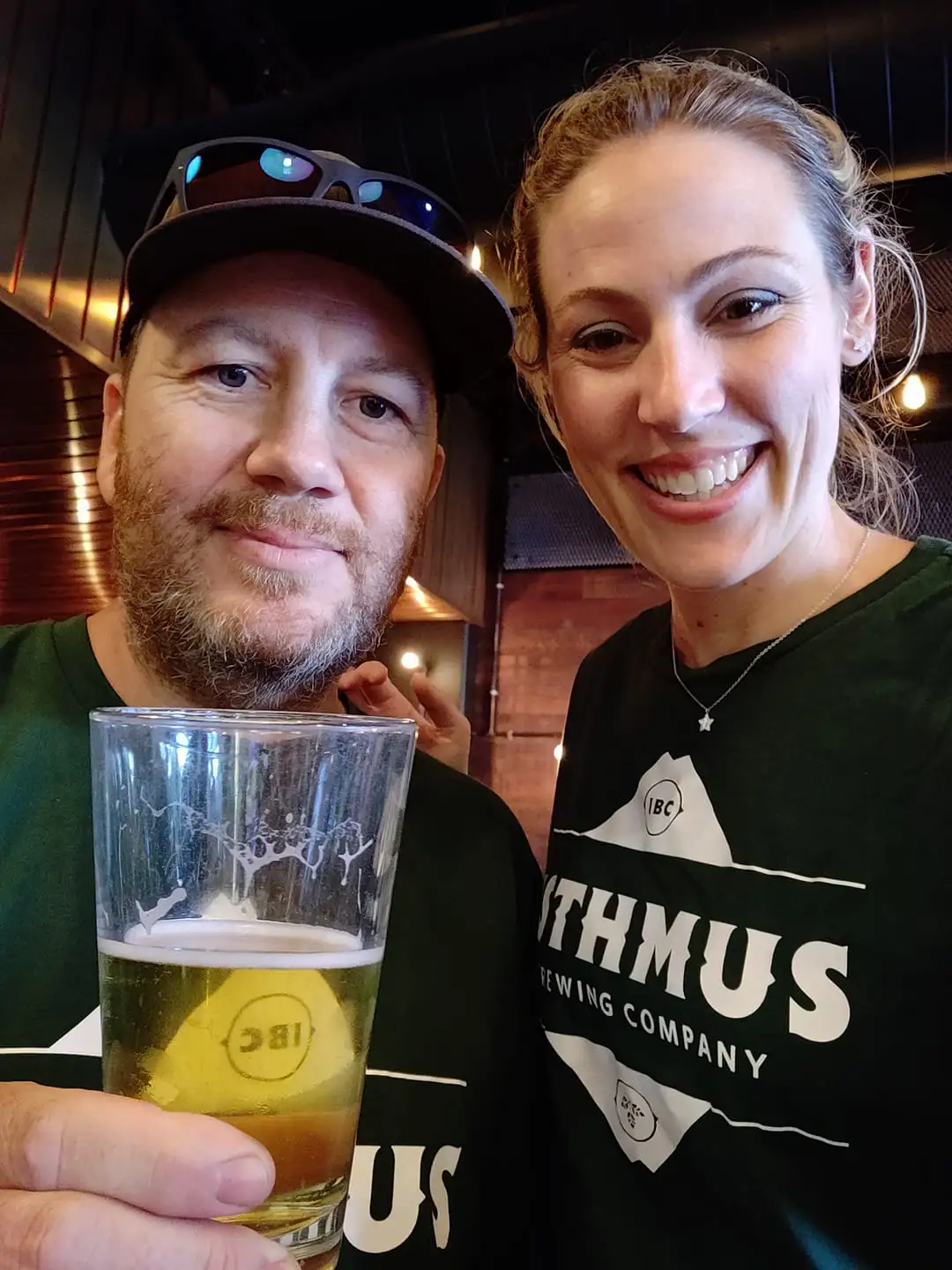 A man holding a pint of beer and a woman stand together, both smiling at the camera. They are indoors, and both wear dark shirts with a brewery logo. The background shows warm lighting and wood decor.