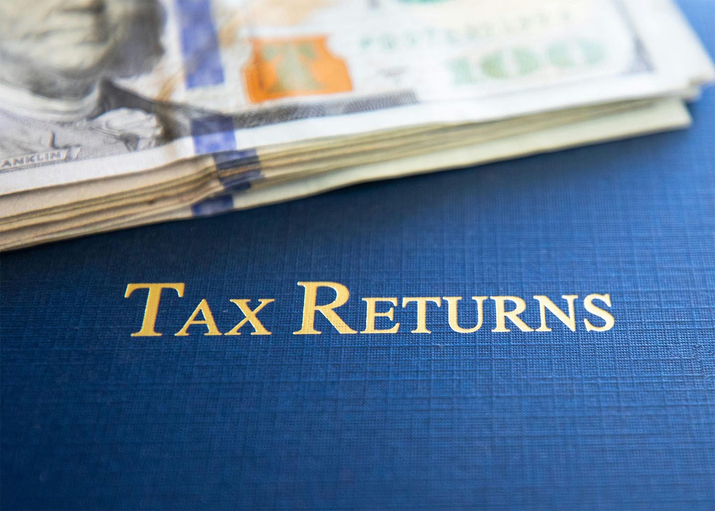 Tax returns document with gold lettering beside a stack of hundred dollar bills on a blue surface