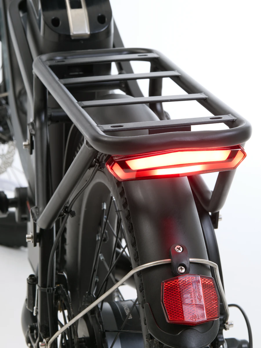 Close-up of the rear section of an electric bicycle, showing the sturdy cargo rack, integrated rear LED light glowing red, tire with fender, and a mounted red reflector for visibility and safety.