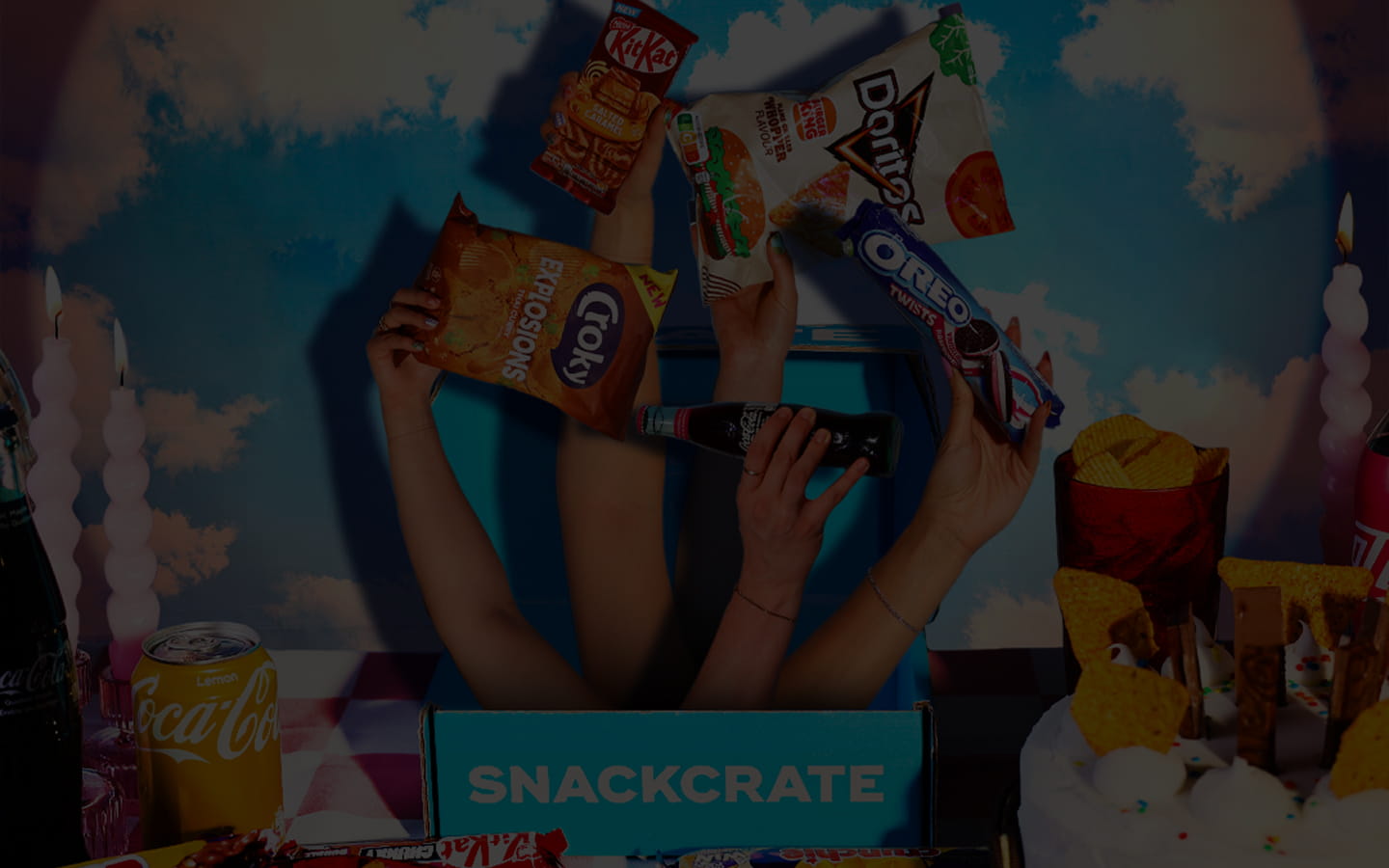 Hands emerging from a blue box labeled 'SNACKCRATE' holding various snacks including Doritos, Oreo Twists, Croky Explosions, and KitKat, with bottles and treats around.