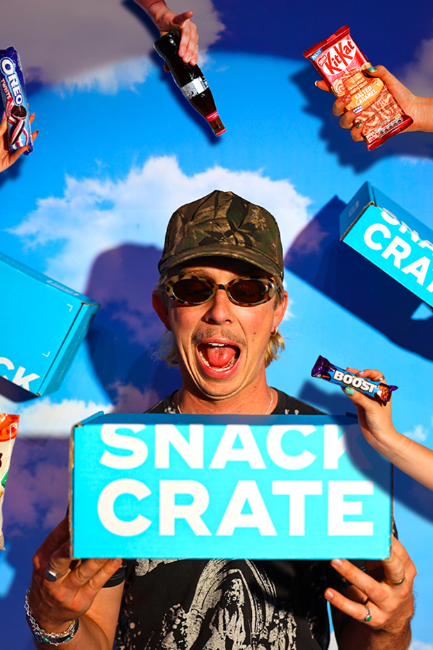 Man wearing a camouflage cap and sunglasses holding a blue Snack Crate box with floating snack items around him against a blue sky background.