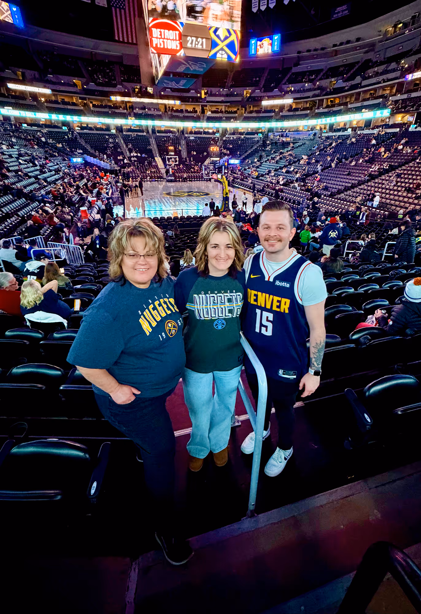 Three Denver Nuggets fans smiling and posing inside an arena with the basketball court and scoreboard showing Detroit Pistons vs Denver Nuggets in the background.