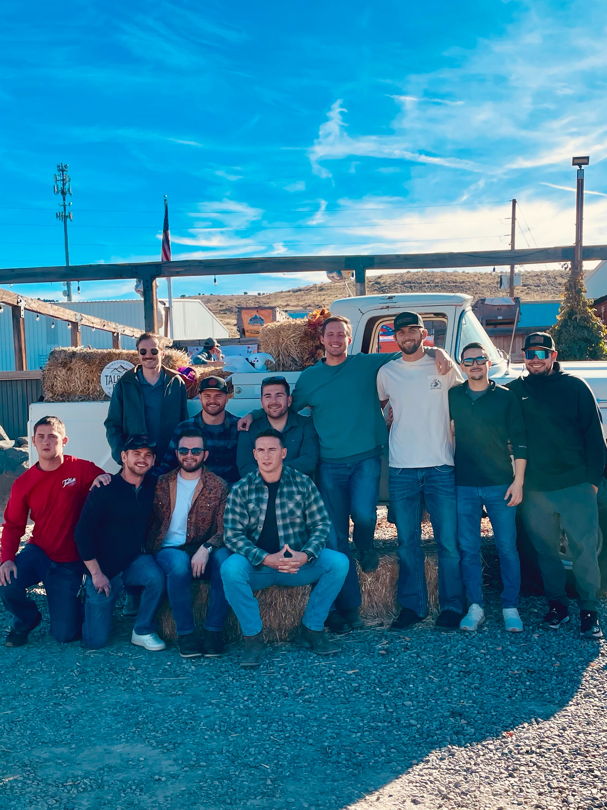 Group of eleven men posing outdoors in front of a white pickup truck with hay bales on a sunny day.