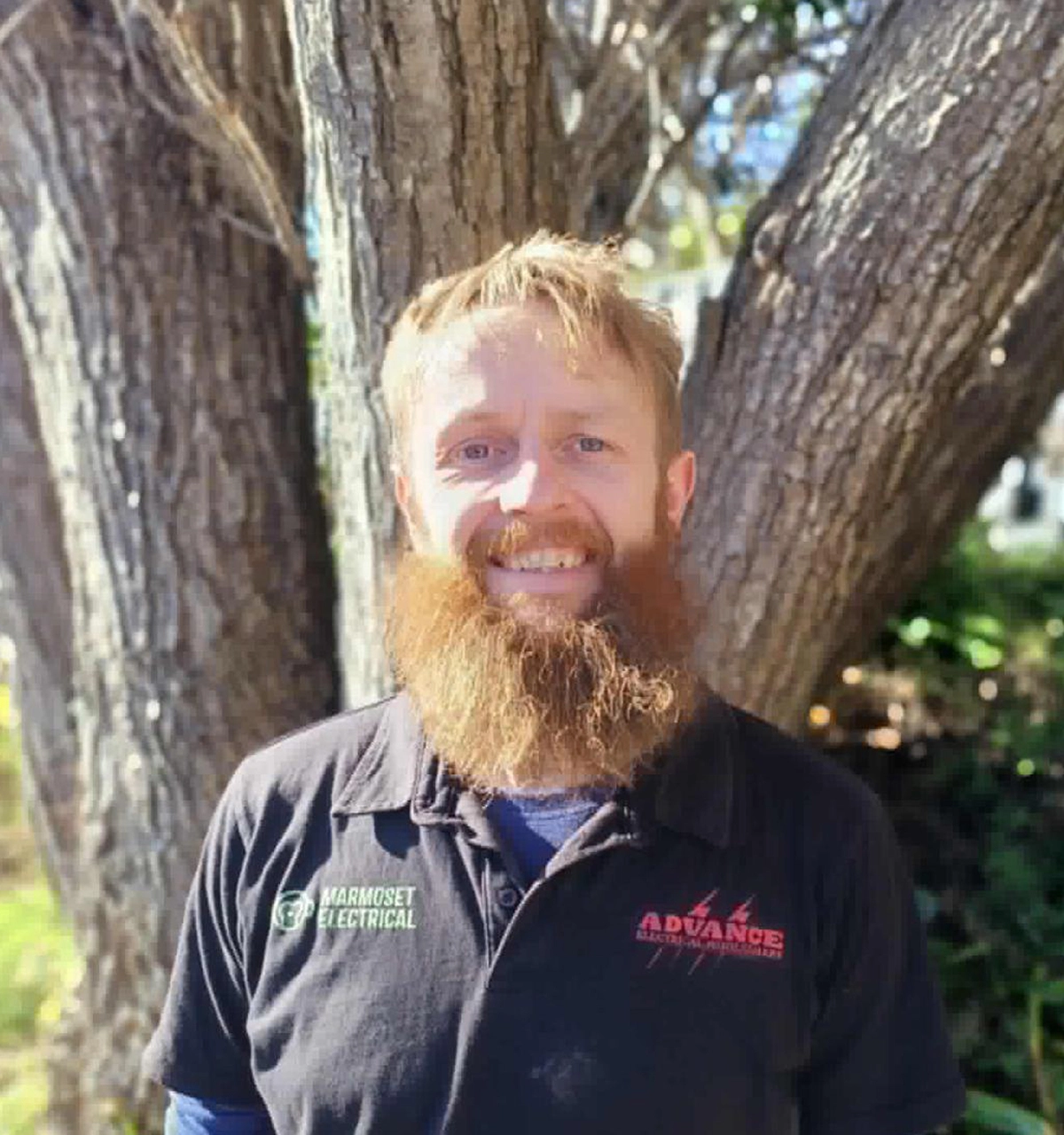 Smiling electrician wearing Marmoset Electrical uniform standing by tree