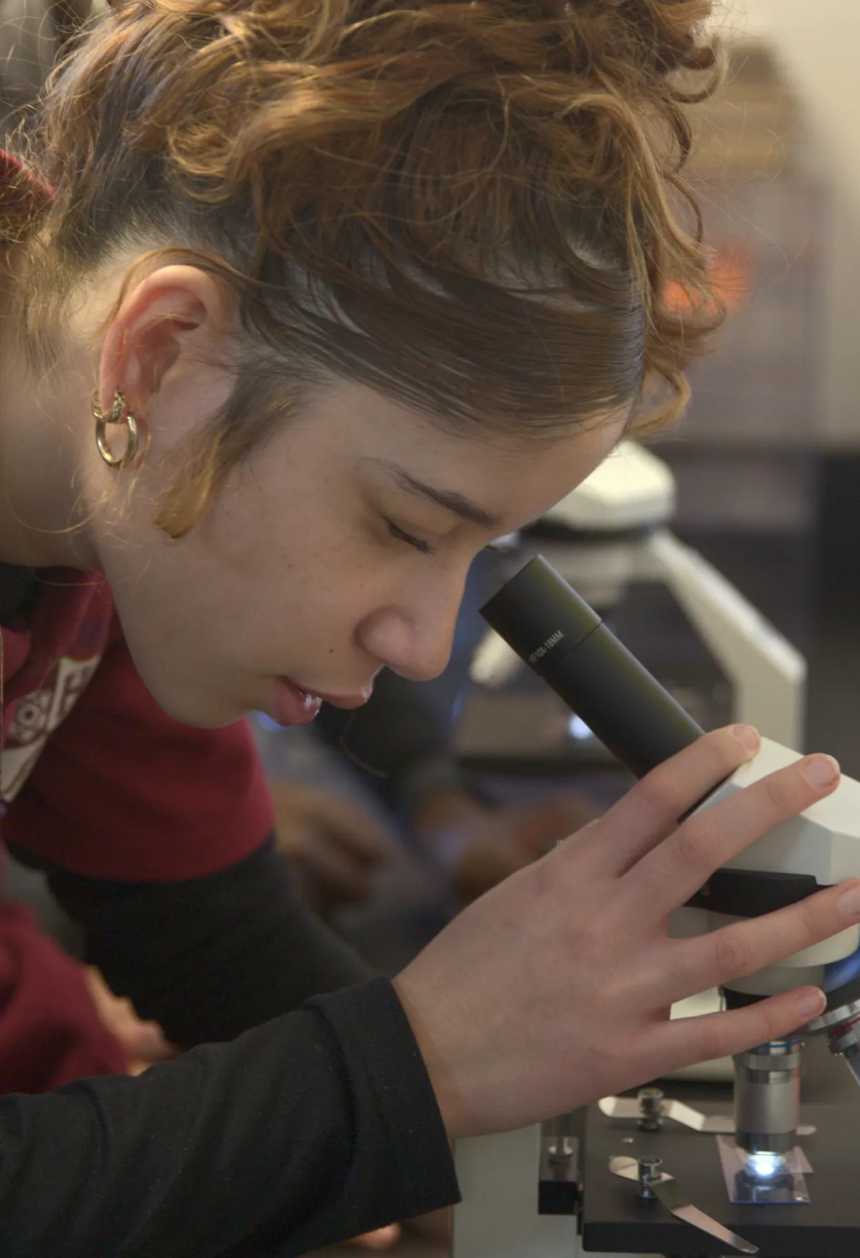 Young woman with curly hair closely examining a slide through a microscope in a science lab.