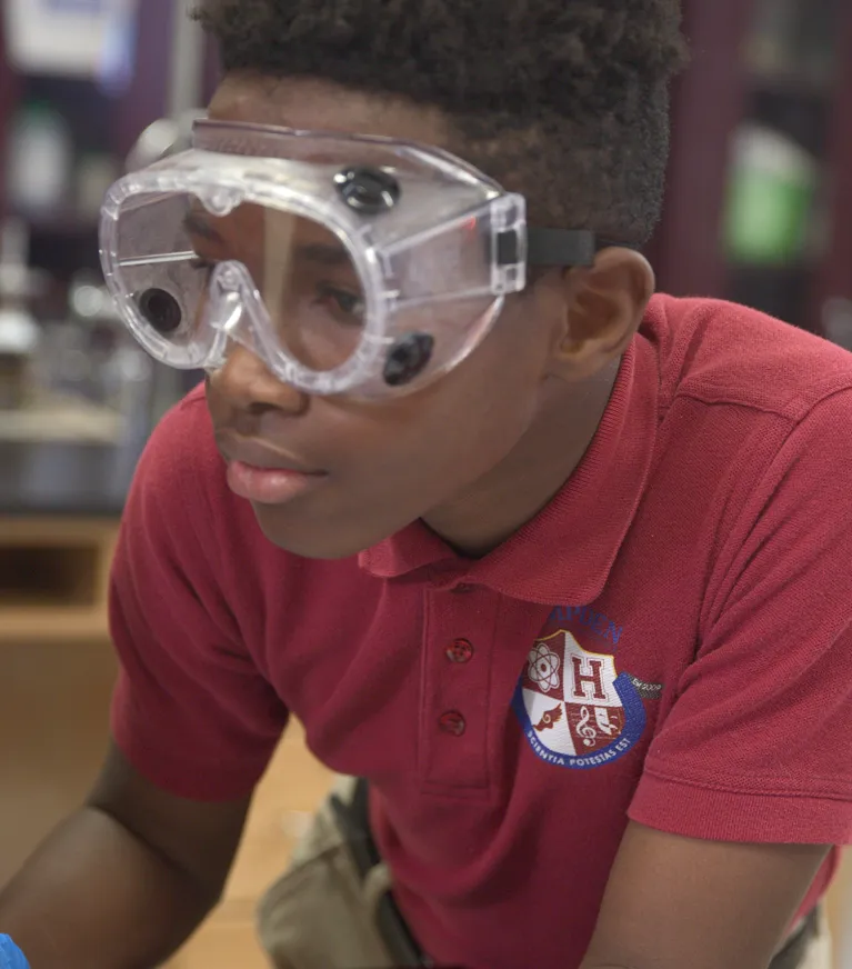 Young student wearing safety goggles and a red polo shirt with a school crest, focused on a science activity.
