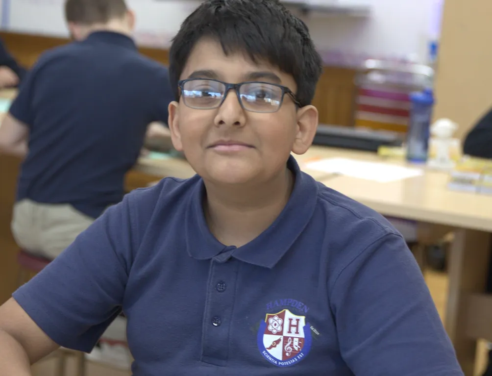 Smiling boy wearing glasses and a navy blue polo shirt with a school emblem sitting in a classroom.