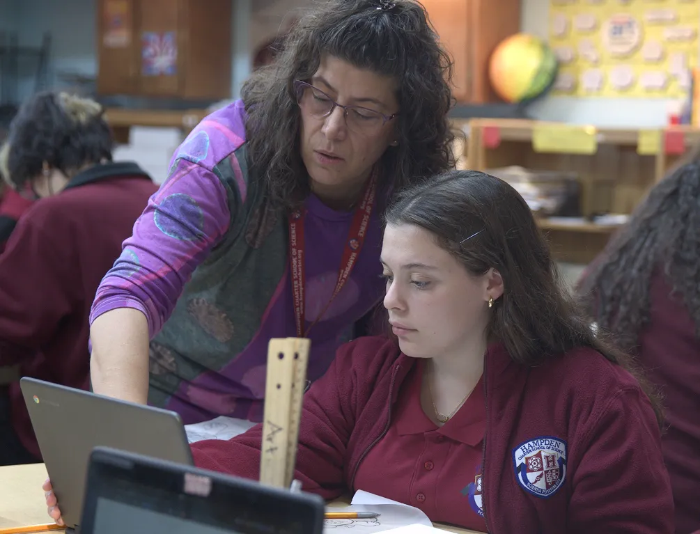 Teacher assisting a female student in a red school uniform with a tablet in a classroom.