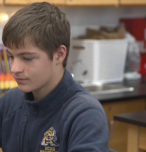 Teenage boy in a navy blue jacket with a tiger logo looking down in a classroom setting.