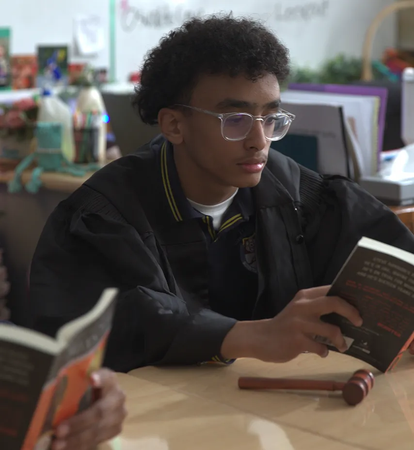 Teenage boy in a judge's robe reading a book at a table with a gavel beside him.
