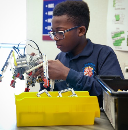 Boy wearing glasses assembling a robotic structure using Lego robotics parts at a desk with yellow and black storage bins.