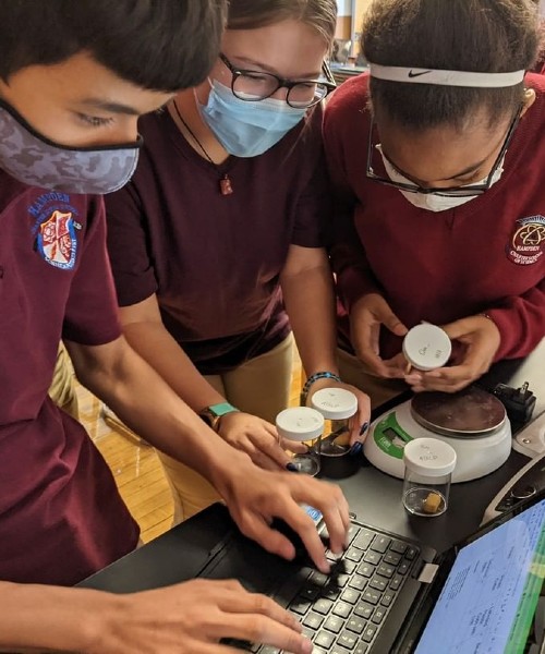 Three students wearing masks and school uniforms engaged in a science experiment using a laptop, small jars, and a digital scale.