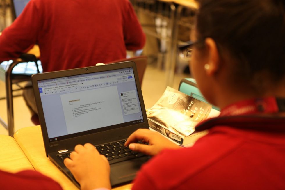 Student wearing red shirt typing on a Lenovo laptop in a classroom with a book titled 'Where She Went' on the desk.