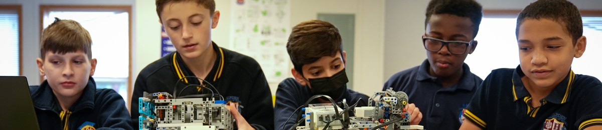 Five boys in school uniforms working on robotics projects at a table indoors.
