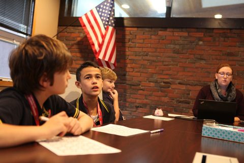 Three boys sitting at a table with papers in front of them, listening to a woman working on a laptop in a room with a brick wall and an American flag.