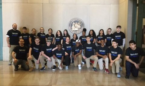 Group of diverse young people and adults wearing matching dark blue shirts posing indoors in front of a wall with a round seal emblem.