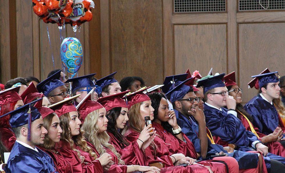 Group of diverse students in red and blue graduation gowns and caps seated indoors during a graduation ceremony.