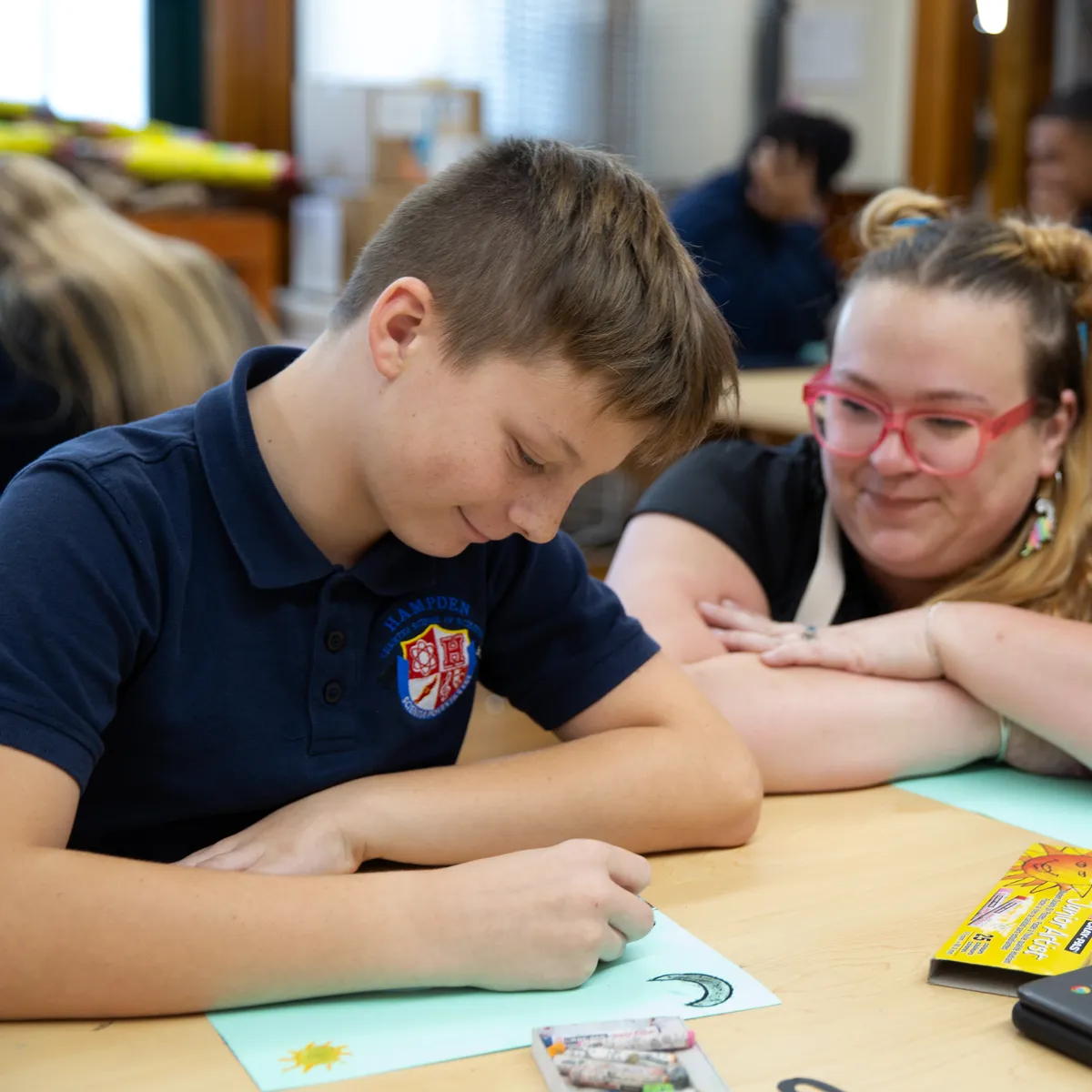 A boy in a navy blue school uniform drawing a crescent moon and sun with crayons while a woman with pink glasses watches him smiling.