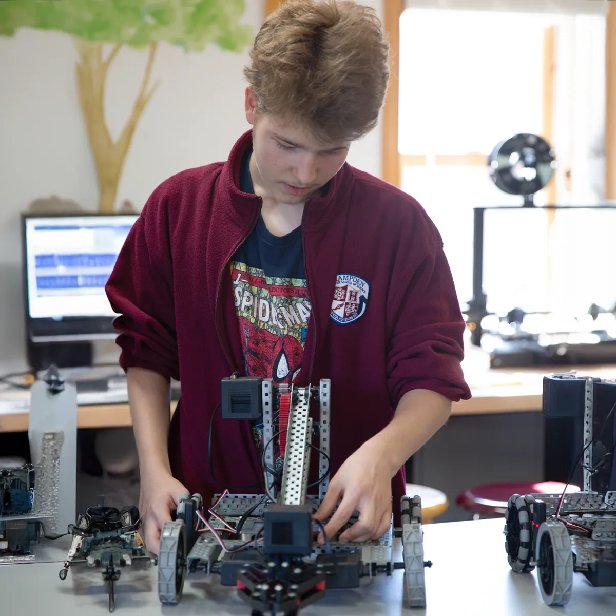 Teenage boy in a maroon jacket assembling a wheeled robot on a table in a workshop.