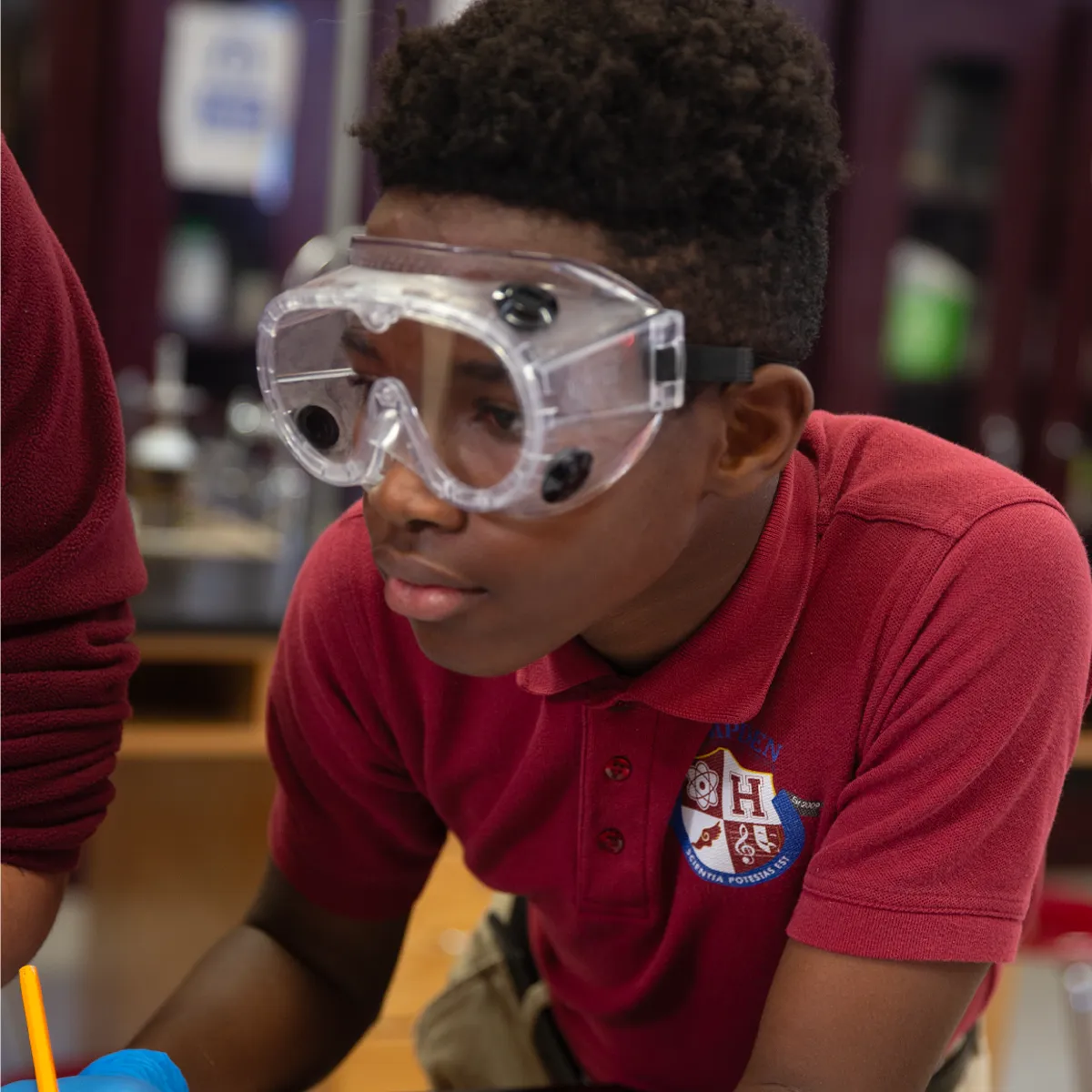 Student wearing clear safety goggles and a red polo shirt with a school emblem, leaning forward in a classroom setting.