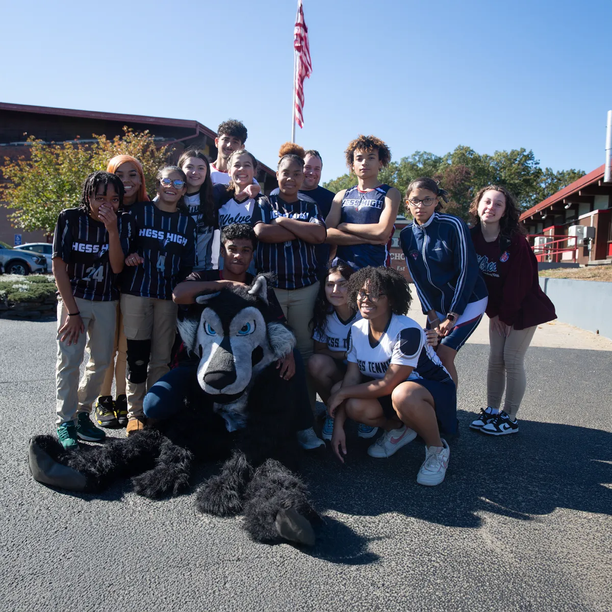 Group of high school students posing outdoors with a wolf mascot in front of a building and American flag.