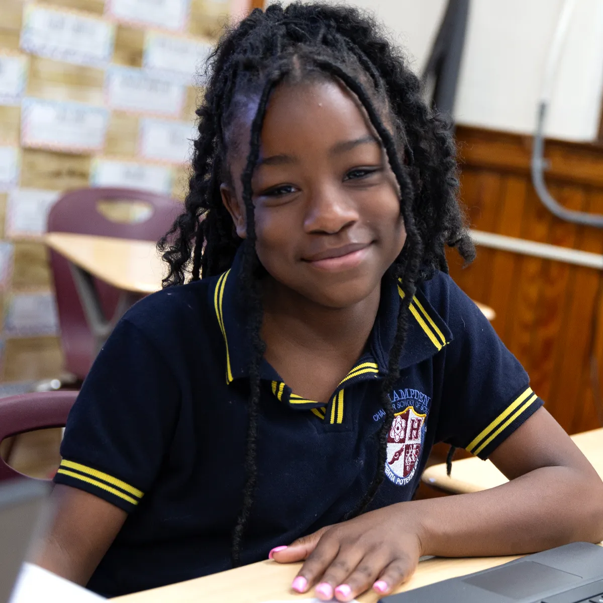 Smiling schoolgirl with braided hair in a navy uniform sitting at a desk in a classroom.