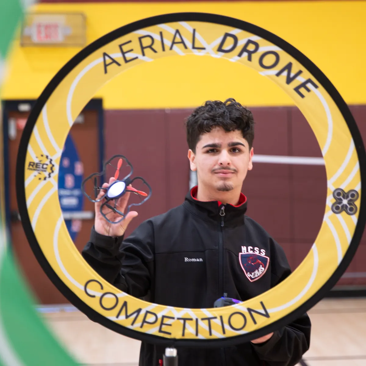 Young man holding a small quadcopter drone in a gymnasium, framed by a yellow ring labeled 'Aerial Drone Competition'.