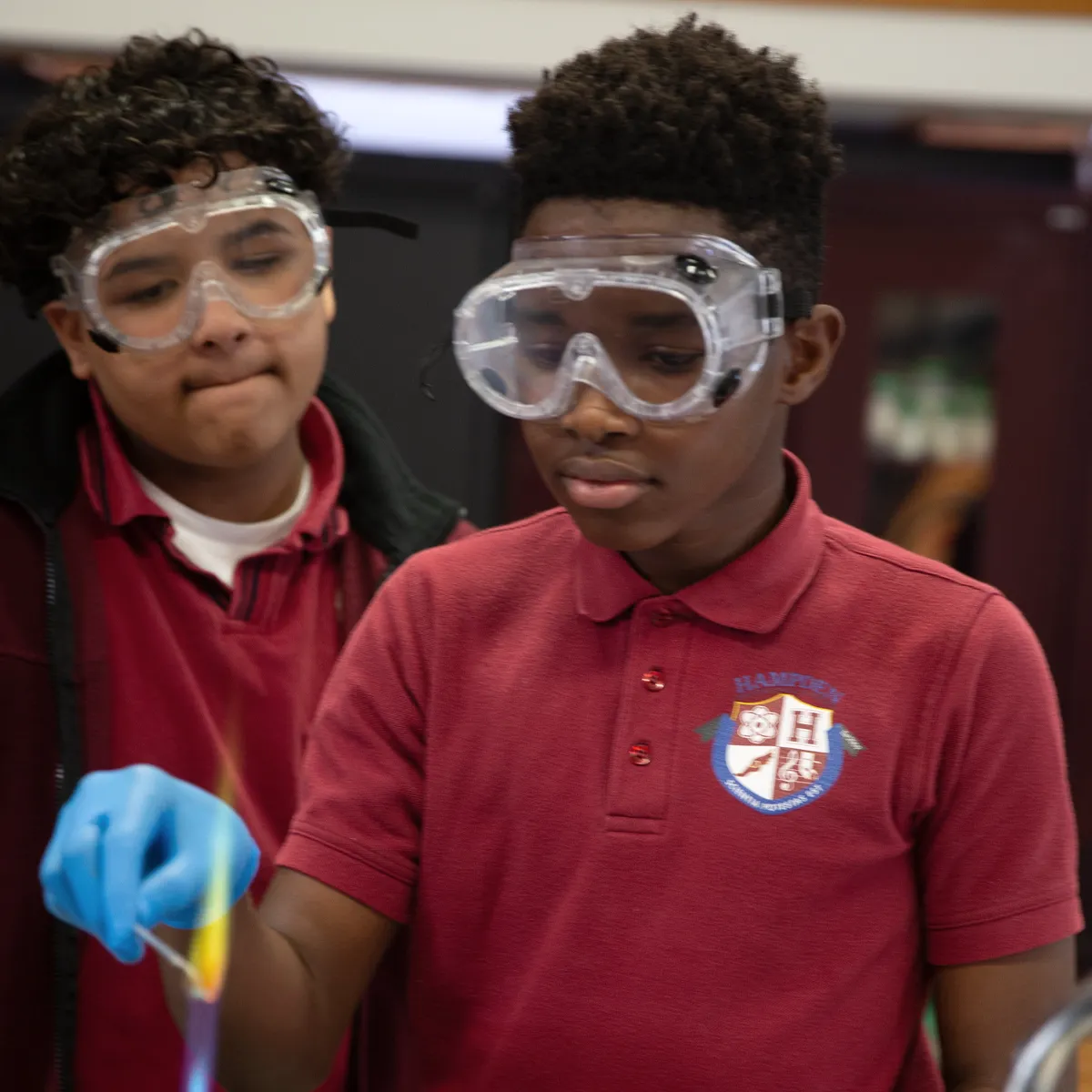 Two students wearing safety goggles and red school uniforms conducting a science experiment with a blue-gloved hand holding a burning material.