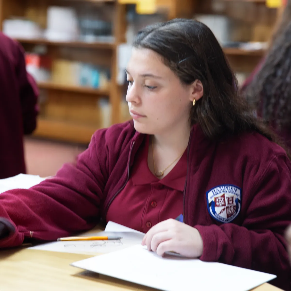 Student wearing a maroon jacket with a school emblem, sitting at a desk with papers and pencils, looking to the side.