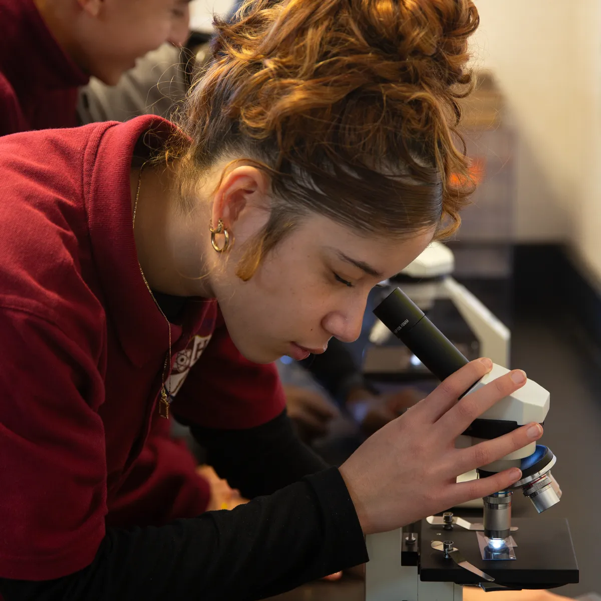 Young woman with curly hair in a maroon shirt looking into a microscope in a lab setting.