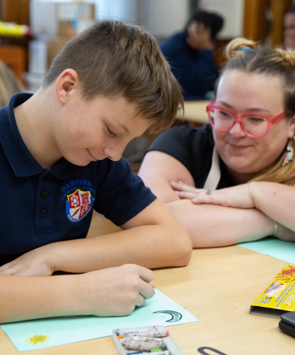 Student drawing a crescent moon on paper while a teacher watches with a smile at a classroom table.