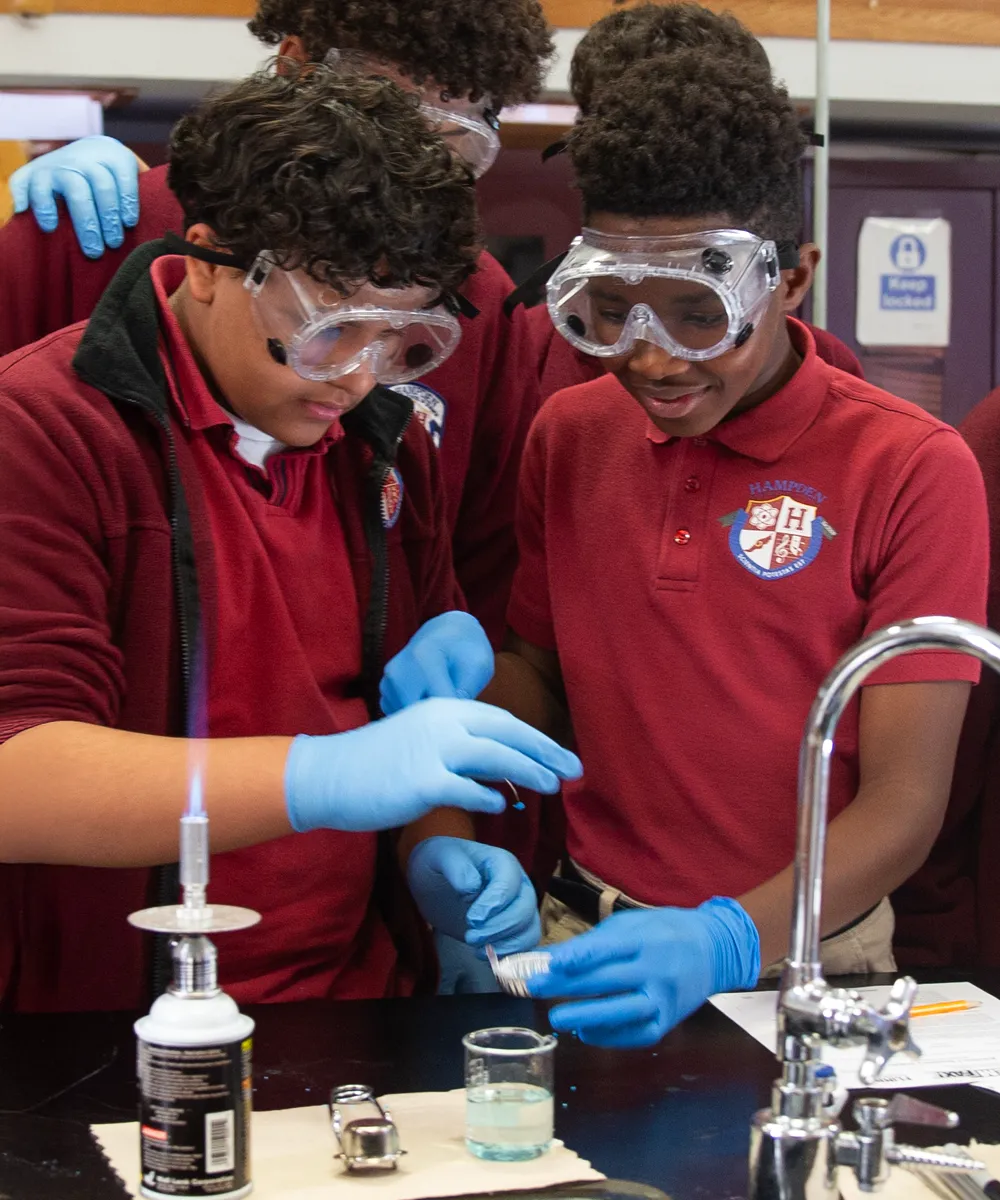 Two students wearing protective goggles and gloves conducting a science experiment with a lit Bunsen burner and glass beaker in a lab.
