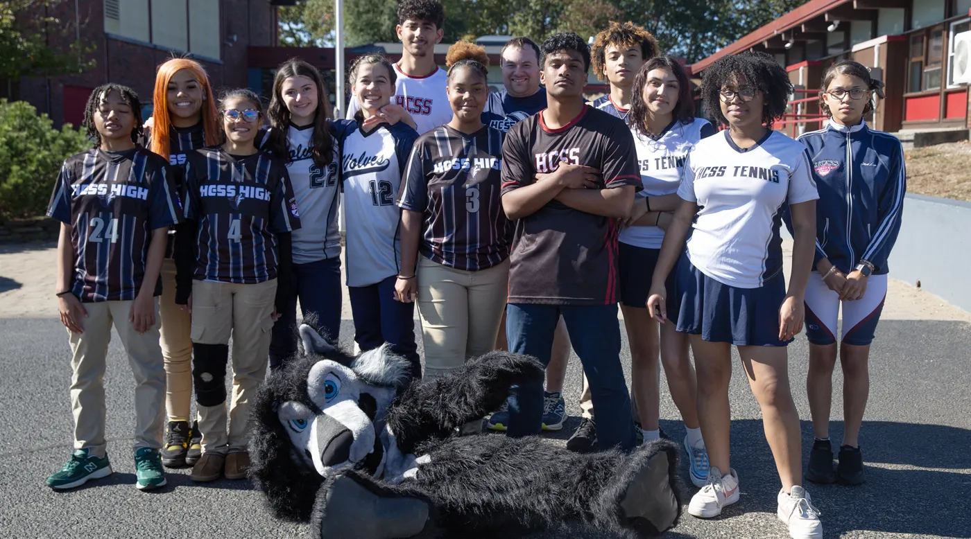 Diverse group of high school students in sports uniforms posing outdoors with a wolf mascot costume lying in front.