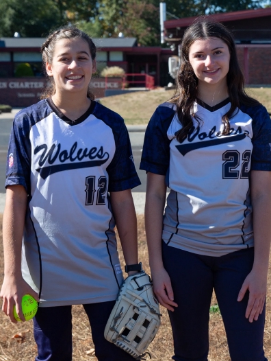 Two female softball players in Wolves uniforms standing on a field, one holding a softball and glove.