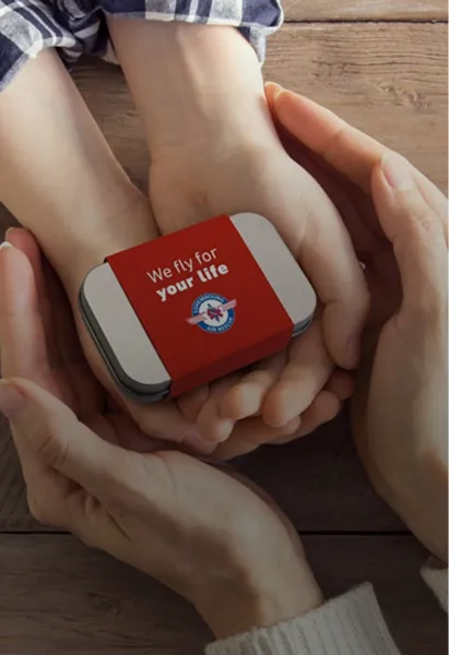 Hände eines Erwachsenen, die sanft die Hände eines Kindes halten, auf denen eine kleine Metallbox mit einem roten Band mit der Aufschrift „Wir fliegen um dein Leben“ liegt.