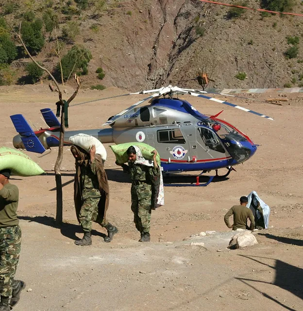 Militärpersonal trägt Säcke auf den Schultern in der Nähe eines geparkten grau-blauen Hubschraubers in einer trockenen, bergigen Gegend.