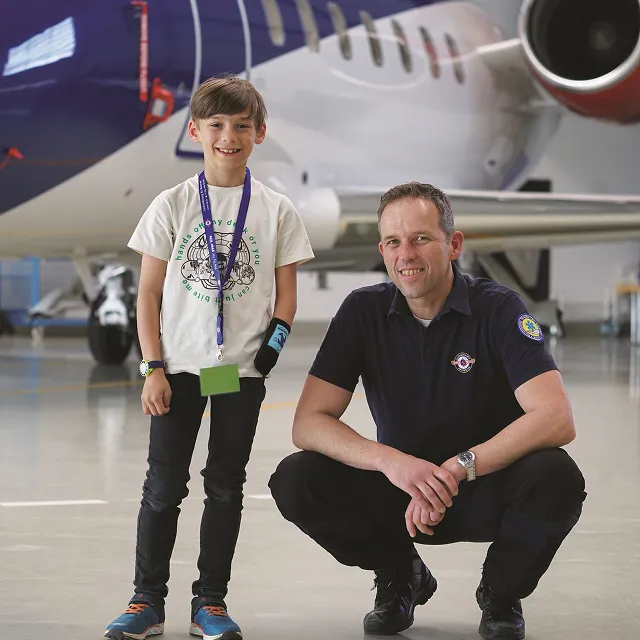 Smiling boy standing next to a squatting man in a hangar with a private jet in the background.