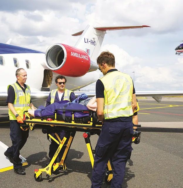 Ärzteteam in Warnwesten schiebt einen Patienten auf einer Trage in Richtung eines rot-weißen Ambulanzflugzeugs auf einem Rollfeld des Flughafens.