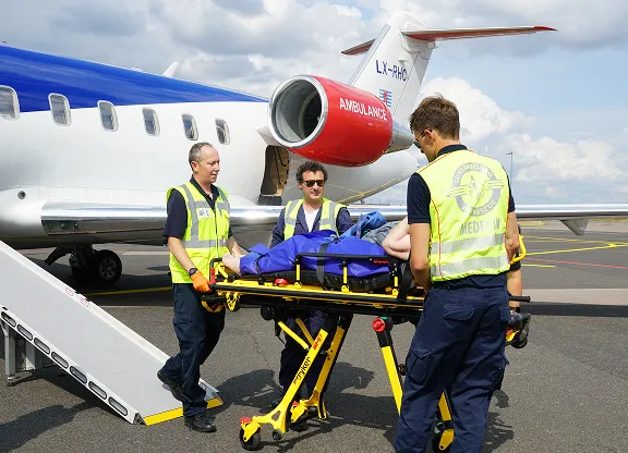Drei Sanitäter in gelben Westen laden einen Patienten auf einer Trage in ein Ambulanzflugzeug auf dem Rollfeld.